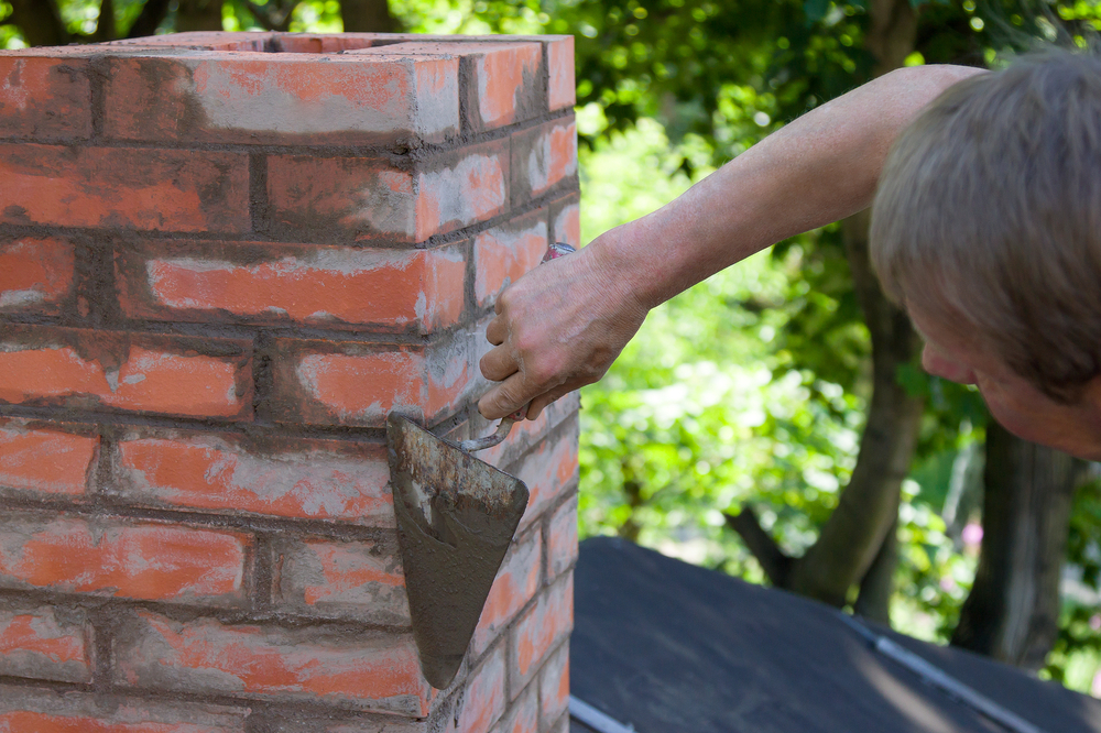 A person uses a trowel to apply mortar to the joints of a brick chimney outdoors, performing chimney repair in Norfolk and Plymouth County, MA, with green trees visible in the background.