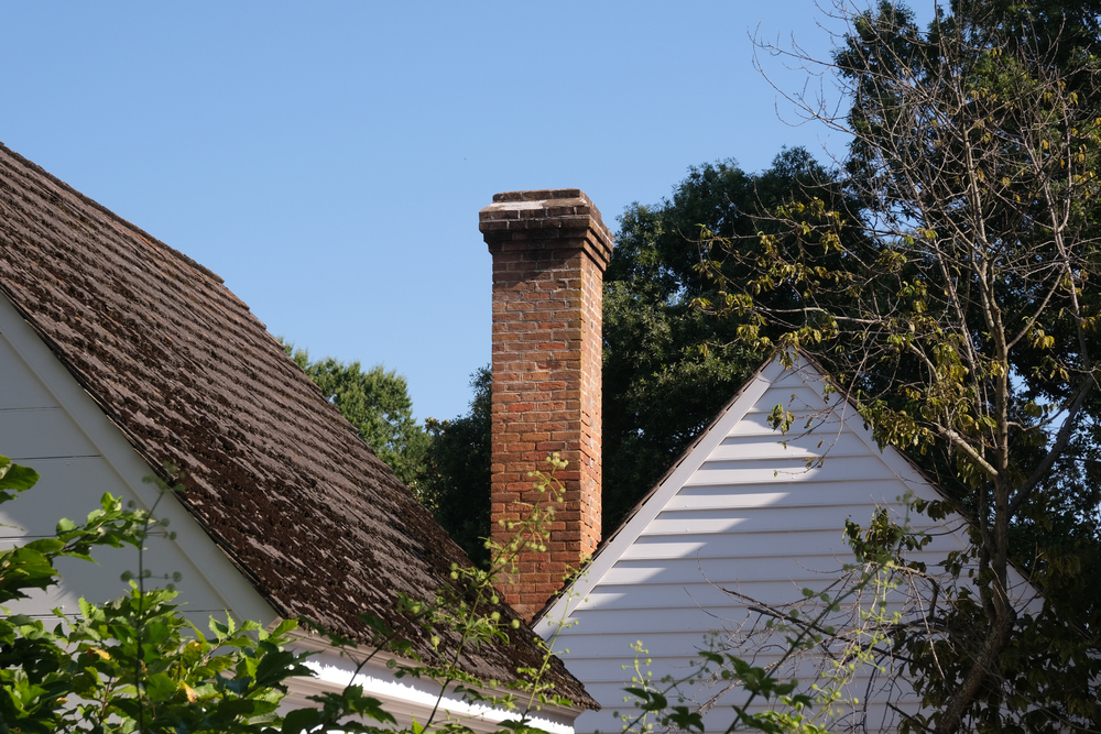 A red brick chimney rises between two white house rooftops, one with wooden shingles, in a charming MA neighborhood. Green trees and leafy branches surround the scene under a clear blue sky.