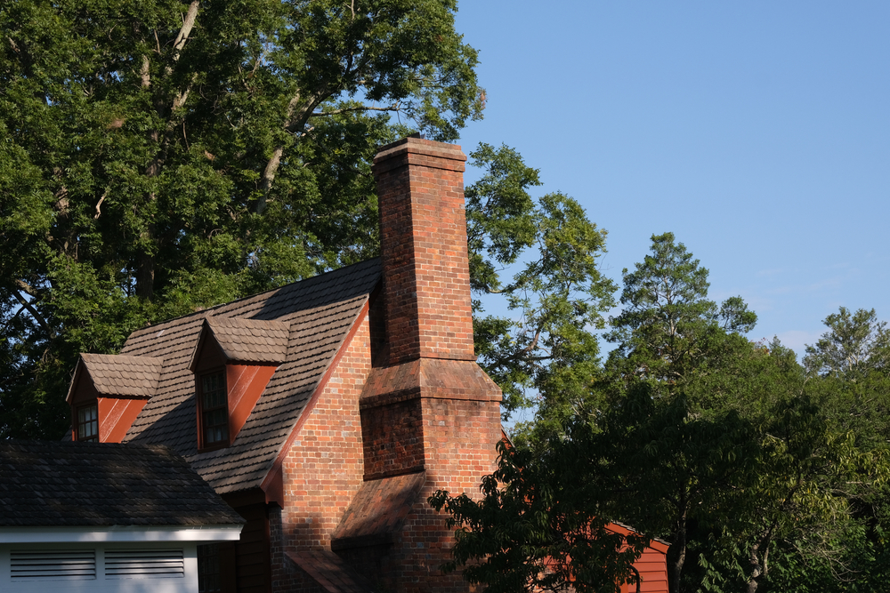 A brick chimney rises above a house with a shingled roof and dormer windows, surrounded by green trees under a clear MA sky—perfect inspiration for chimney repair in Norfolk and Plymouth County.