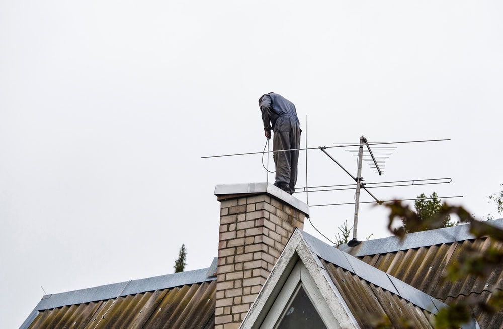 A person stands on a sloped roof in MA, working on chimney repair in Norfolk and Plymouth County. Tools are in hand as they work on the brick chimney near an antenna, set against a cloudy sky. The person's face is not visible.