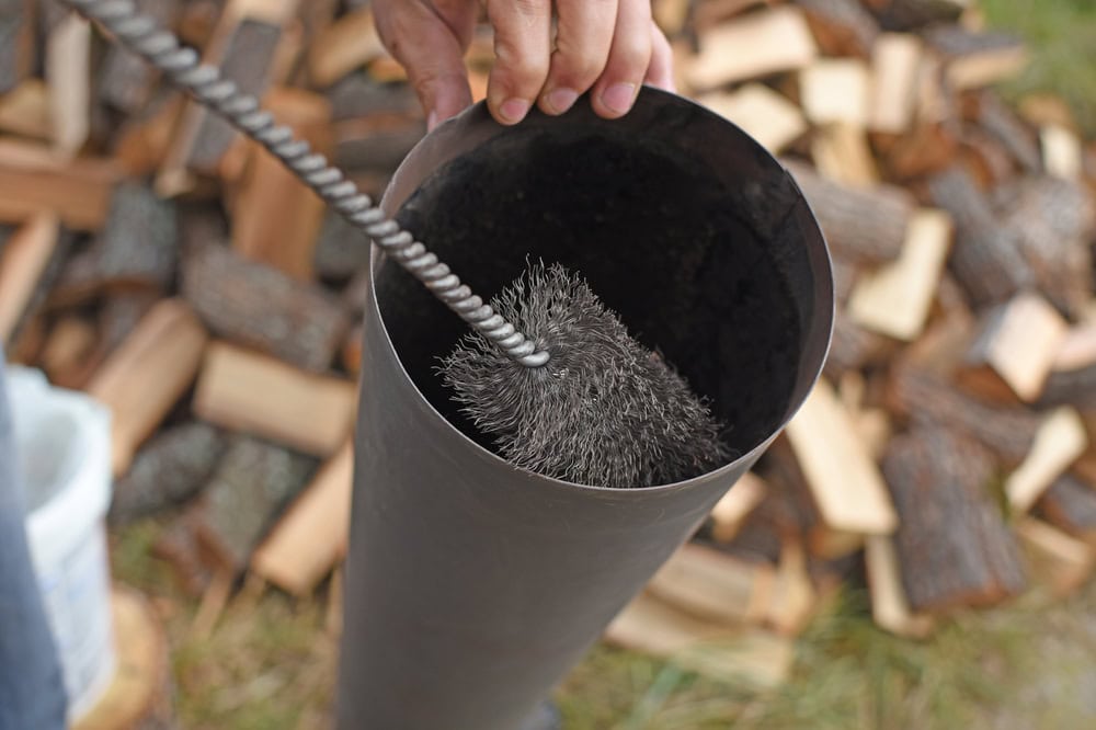 A hand holds a chimney brush inside a metal stove pipe, preparing to clean it. Chopped firewood is scattered on the ground in the background—a typical scene for a chimney sweep in Norfolk and Plymouth County, MA.