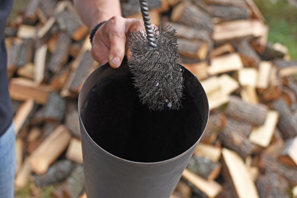 A person cleans the inside of a metal chimney pipe with a wire brush, maintaining safety. In the background, stacked pieces of cut firewood are visible—a scene common for chimney sweep Norfolk and Plymouth County, MA services.