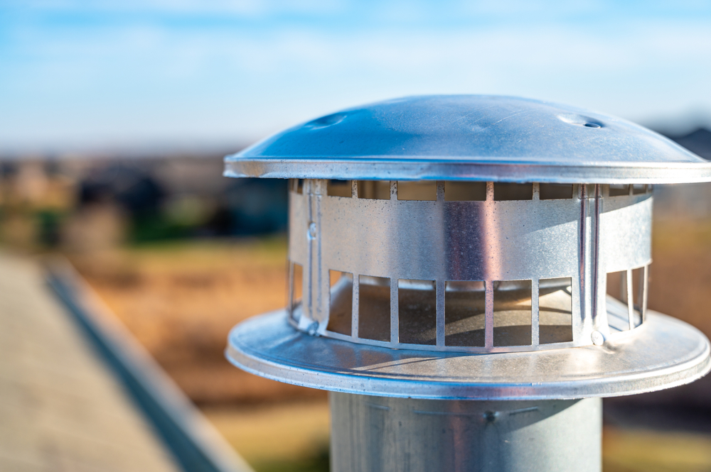 Close-up of a shiny metal chimney cap on a rooftop, highlighting expert chimney repair Norfolk and Plymouth County, with houses and open landscape softly blurred under a bright blue sky.
