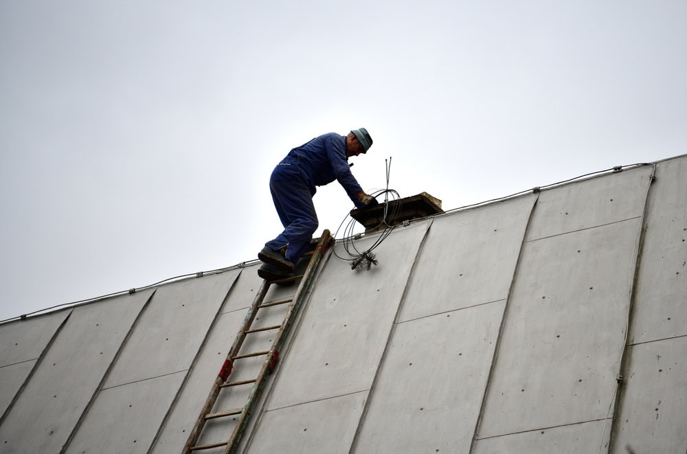 A worker in blue overalls and a helmet stands on a ladder propped against a sloped concrete roof, handling electrical wires near a vent on a cloudy day—perfect for chimney repair Norfolk and Plymouth County, MA.