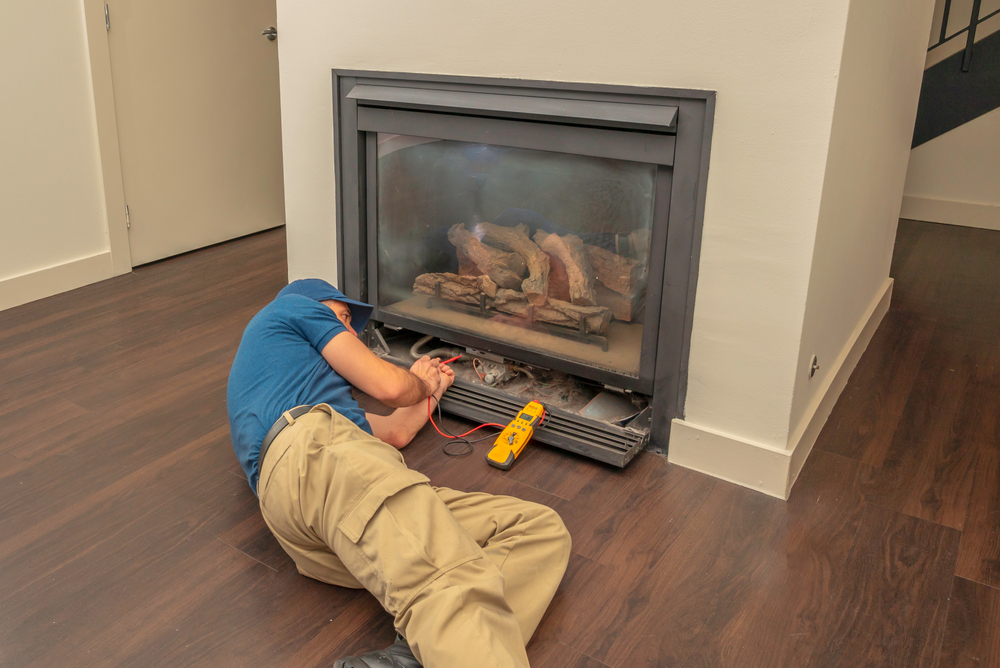 A person in a blue shirt and khaki pants lies on the floor while working on a gas fireplace, tools and a digital multimeter nearby—perfect for those needing chimney repair in Norfolk and Plymouth County.