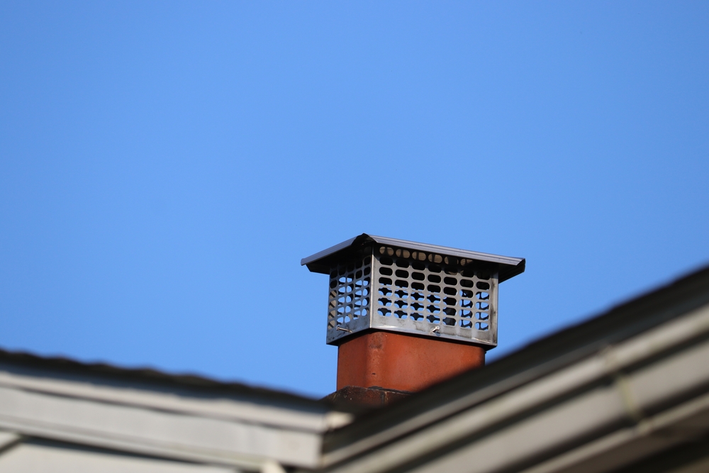 A metal chimney cap sits atop a brick chimney on a house in MA, possibly serviced by a chimney sweep Norfolk and Plymouth County,MA. The roof edges frame the bottom of the image, set against a clear blue sky.