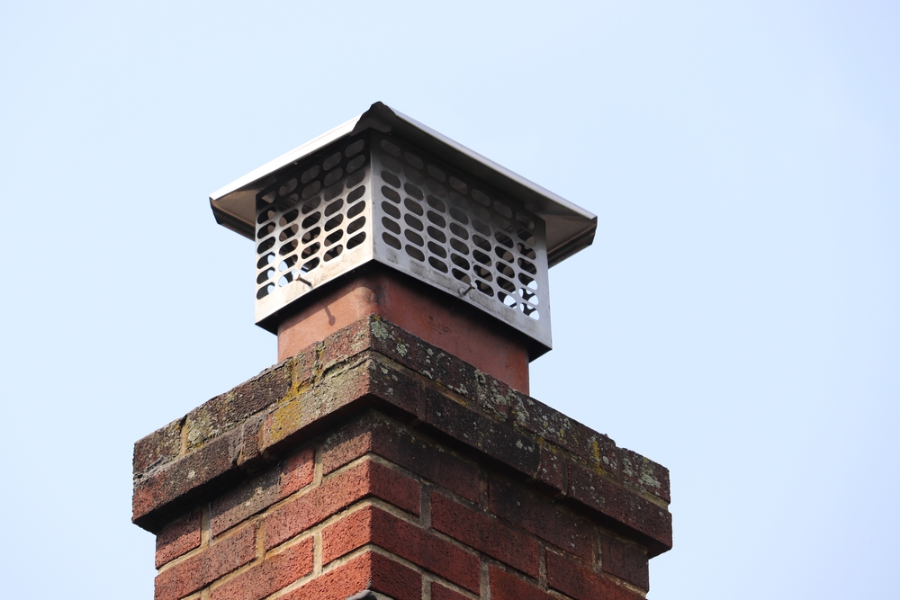 A close-up of a brick chimney with a metal cap and mesh cover on top, set against a clear blue sky—typical of homes serviced by chimney sweep Norfolk and Plymouth County, MA.