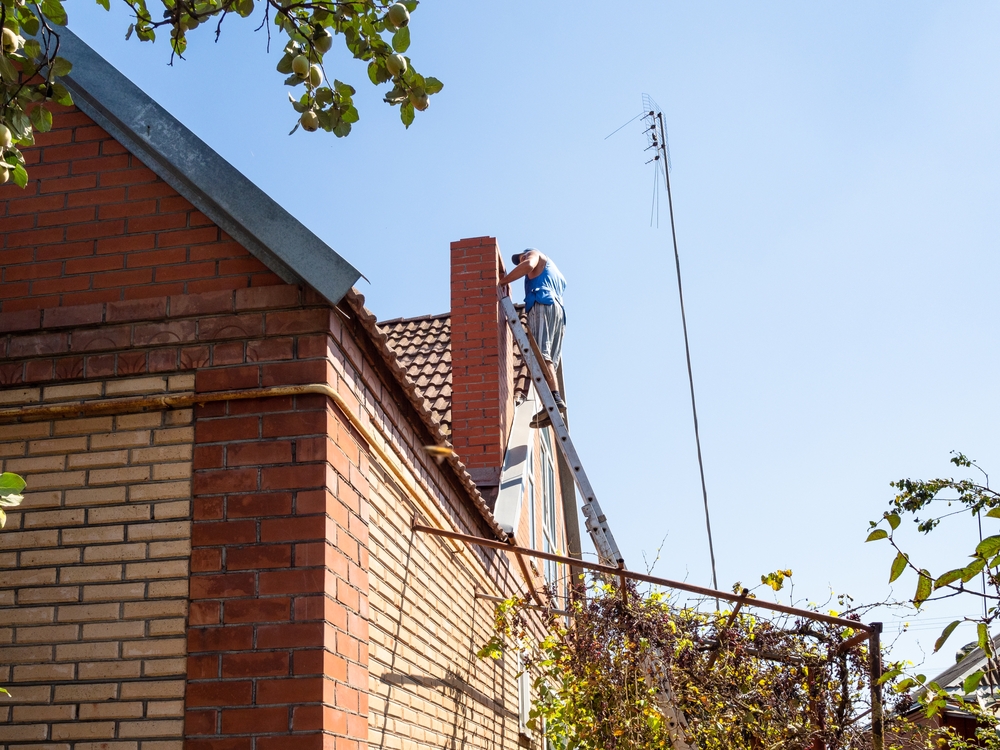 A person stands near the top of a tall ladder, working on a brick chimney on the roof of an MA house, with clear blue sky and tree branches in the background.