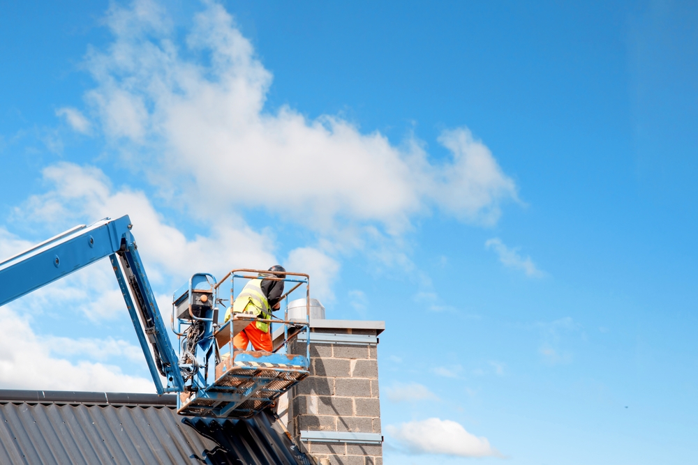 A worker in a high-visibility vest and helmet stands on an elevated platform next to a brick chimney, performing chimney repair in Norfolk and Plymouth County, MA, on a rooftop under a blue sky with scattered clouds.