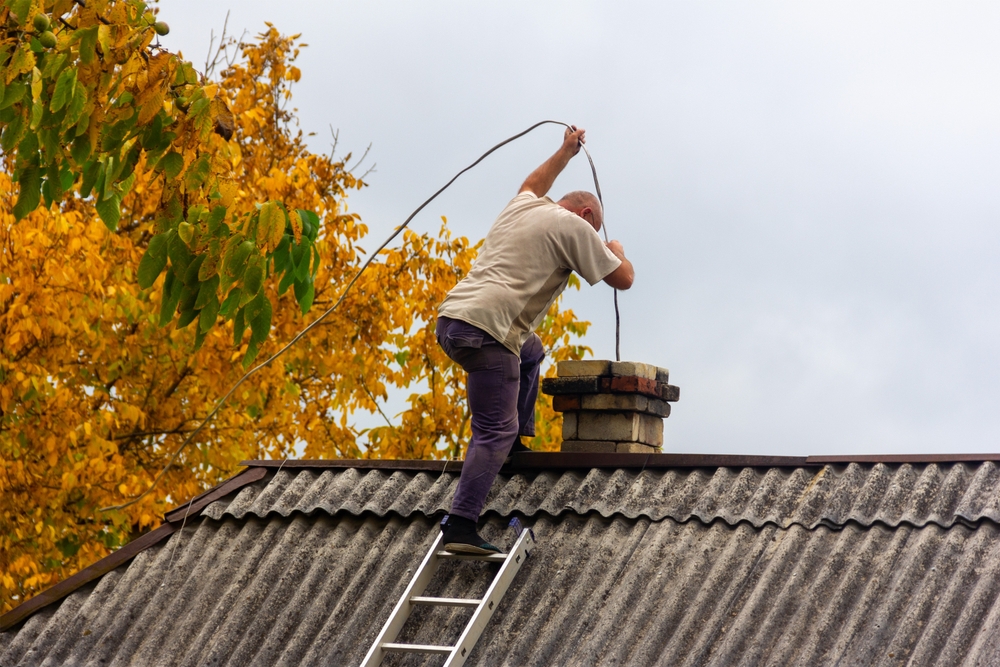 A person standing on a sloped roof next to a chimney, holding a long wire or rope, with a ladder propped against the roof—perfectly capturing chimney repair in Norfolk and Plymouth County, MA, with autumn-colored trees in the background.