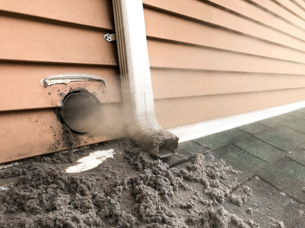 A dryer vent on the side of a house with tan siding is releasing lint and dust onto the roof, a sign of a clogged vent. Nearby, a white gutter downspout runs vertically—chimney restoration Norfolk and Plymouth County, MA services are available.