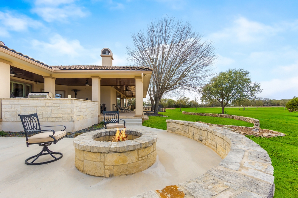 A cozy backyard patio with stone seating surrounds a fire pit, perfect for relaxing after a chimney sweep in Norfolk and Plymouth County. Two chairs face the fire, while the patio connects to a house with a covered porch and views of green lawn and trees.