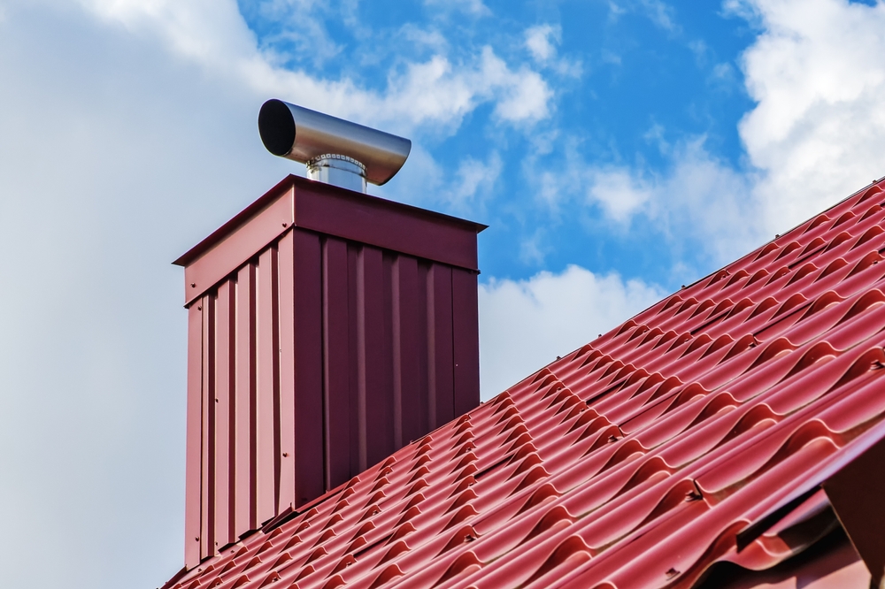 A red metal chimney with a modern cap sits on a sloped roof of red metal tiles, set against a blue sky with white clouds—perfect inspiration for chimney restoration in Norfolk and Plymouth County, MA.