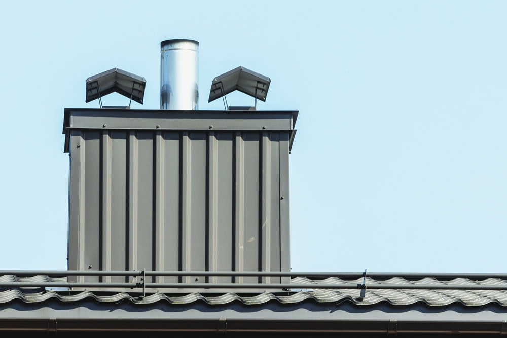 A close-up view of a modern metal chimney on a rooftop in MA, featuring two black vent covers and a shiny metal flue pipe against a clear blue sky—ideal for those seeking a chimney sweep in Norfolk and Plymouth County.
