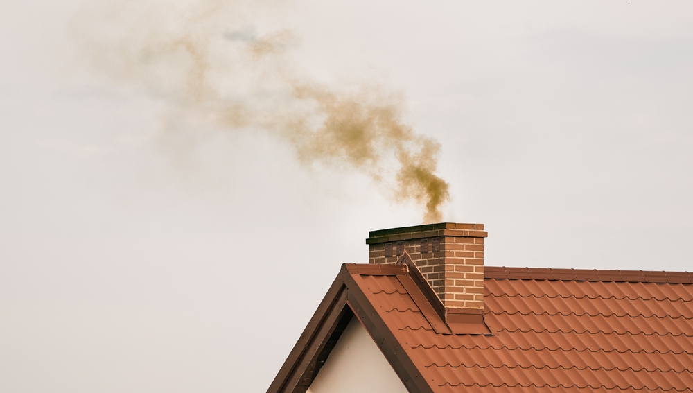 A brick chimney on a house with a red tiled roof in MA releases brownish smoke into a cloudy sky, highlighting the importance of regular chimney sweep Norfolk and Plymouth County services.