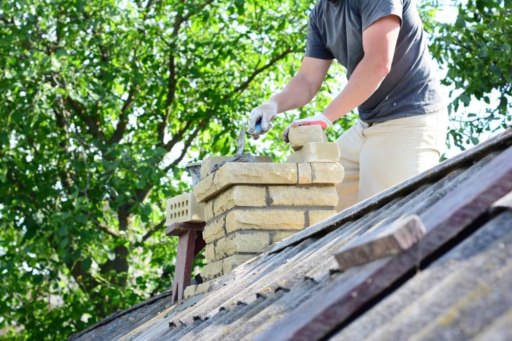 A person building or repairing a brick chimney on a sloped roof with mortar and bricks, showcasing expert chimney repair Norfolk and Plymouth County skills, while green trees are visible in the background.