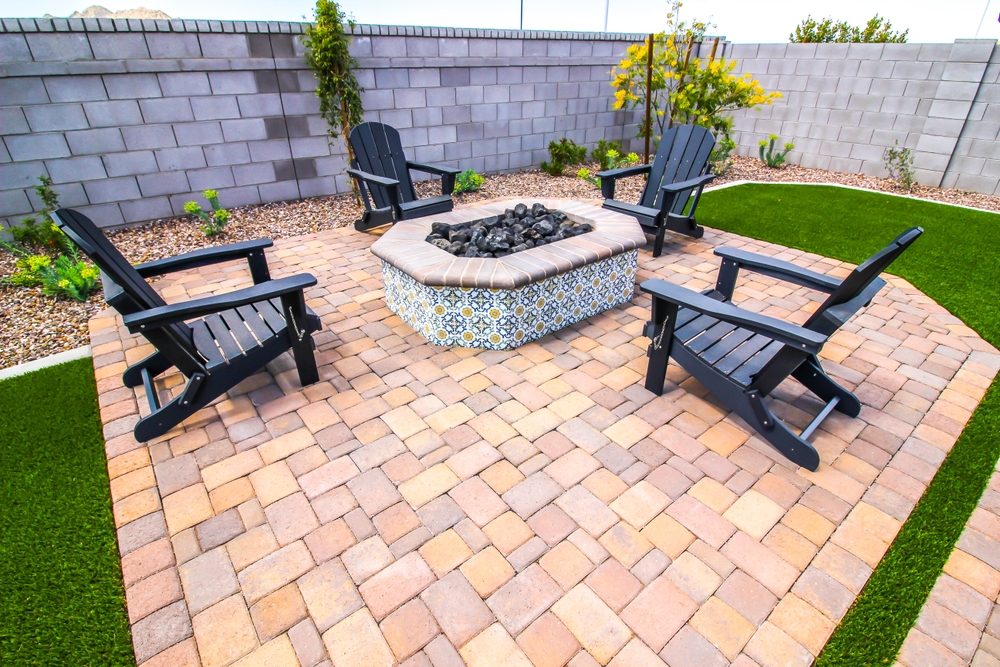 A backyard patio with a tile-trimmed fire pit—ideal after a visit from a chimney sweep in Norfolk and Plymouth County, MA—surrounded by four black Adirondack chairs, tan pavers, and decorative plants along a cinder block fence.