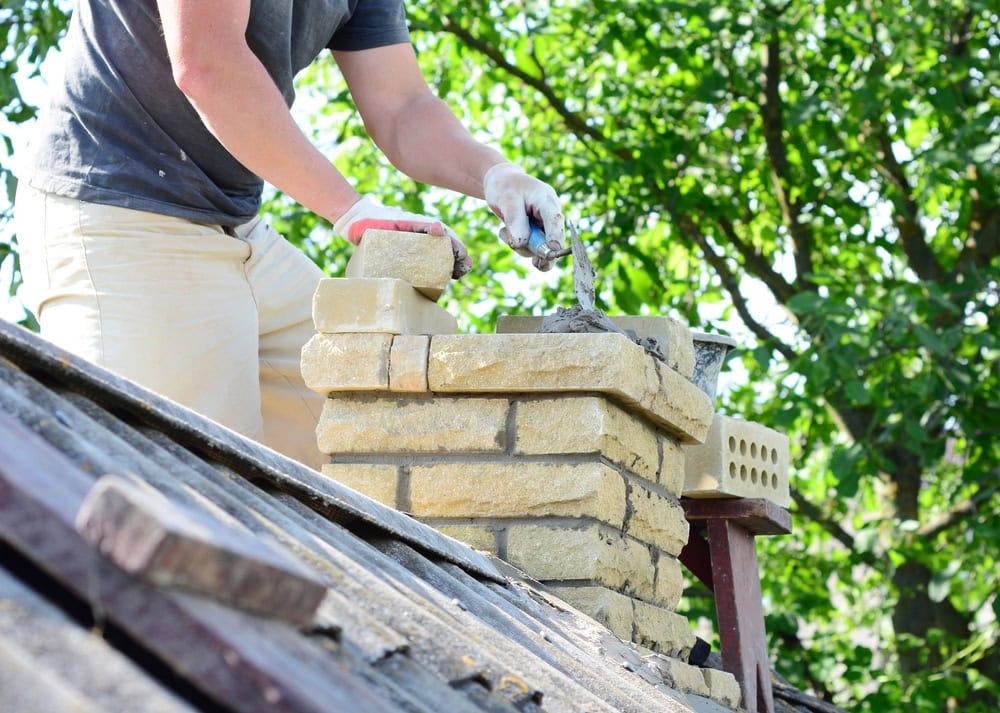 A person in a gray shirt and gloves builds a brick chimney on a roof, using mortar to secure yellow bricks—a typical scene during chimney restoration Norfolk and Plymouth County. Green trees are visible in the background.