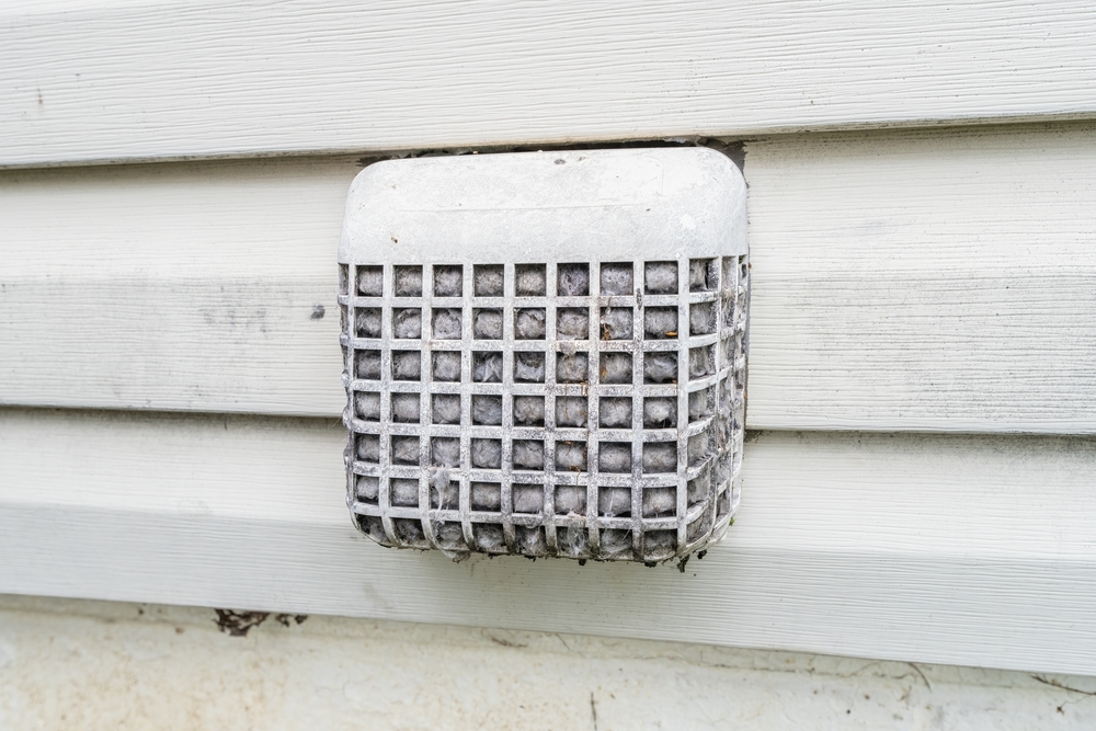 A close-up of a vent on a house exterior with white siding, covered by a mesh screen and heavy lint buildup—much like what chimney sweep Norfolk and Plymouth County experts often find during inspections.