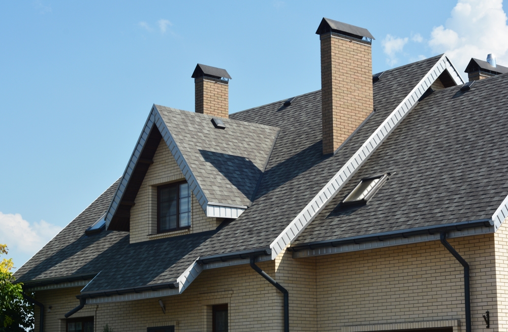 A close-up view of a modern house with yellow brick walls, grey shingle roof, two chimneys—ideal for chimney sweep Norfolk and Plymouth County—an attic window, skylight, and blue sky with light clouds in the background.
