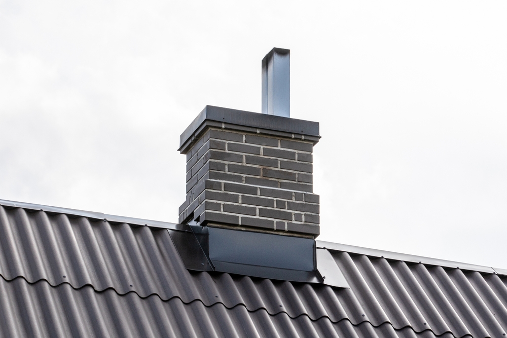 A gray brick chimney with a metal cap rises from a black corrugated metal roof against a cloudy sky, showcasing quality chimney repair in Norfolk and Plymouth County, MA.
