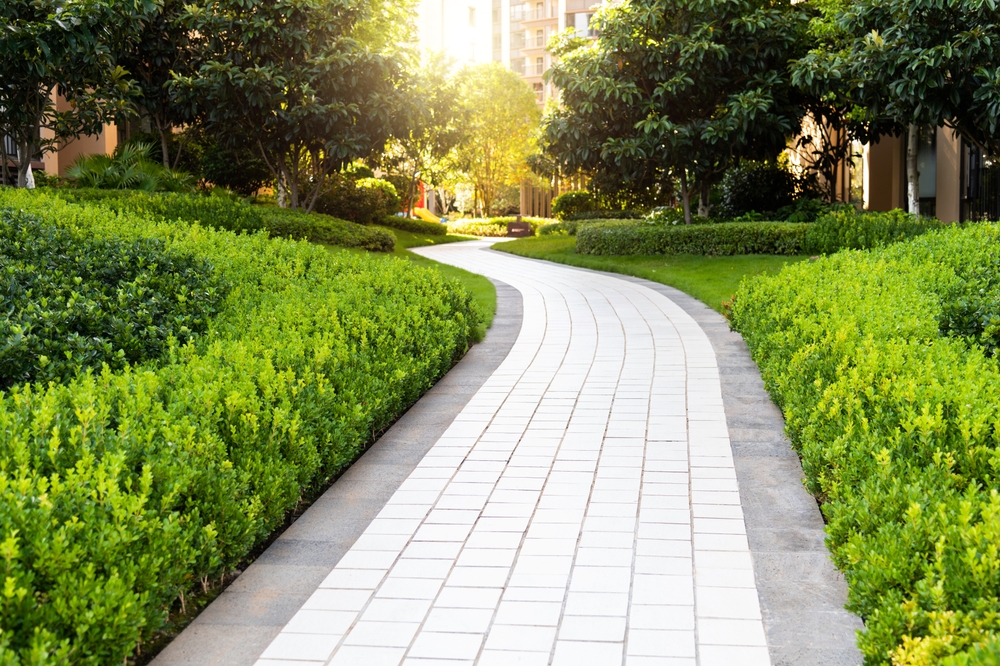 A winding, tiled pathway curves through lush, green gardens with bright shrubbery and trees, leading toward sunlight and apartment buildings in the background—perfect scenery for MA residents seeking expert chimney repair Norfolk and Plymouth County.