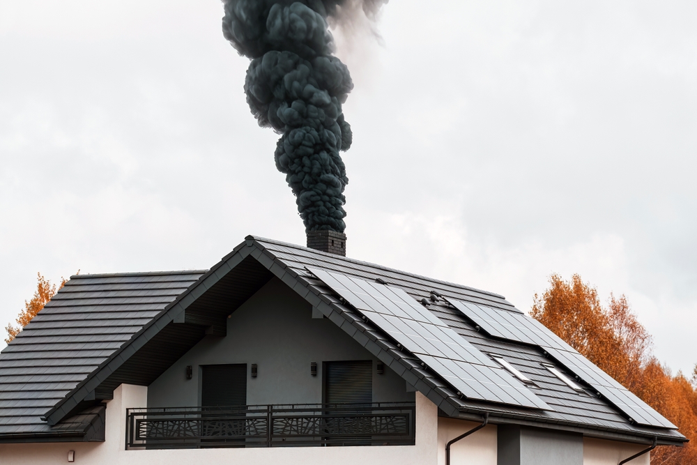 A modern house with solar panels on the roof emits thick black smoke from its chimney, highlighting the need for chimney restoration in Norfolk and Plymouth County, MA. Autumn trees with orange leaves are visible in the background.