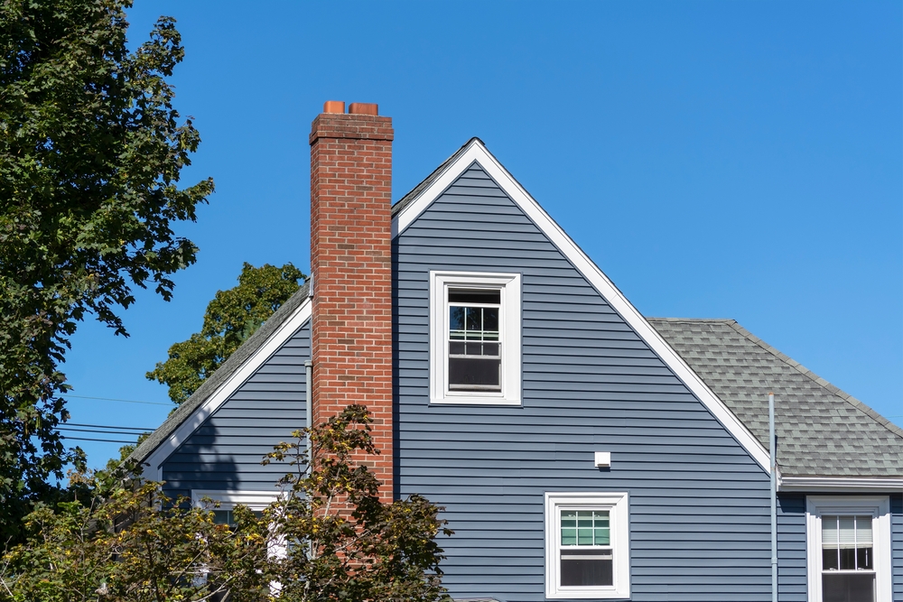 A blue house with white trim and a red brick chimney stands against a clear sky, trees framing its side. Recent chimney repair Norfolk and Plymouth County ensures the home’s classic features remain in excellent condition.