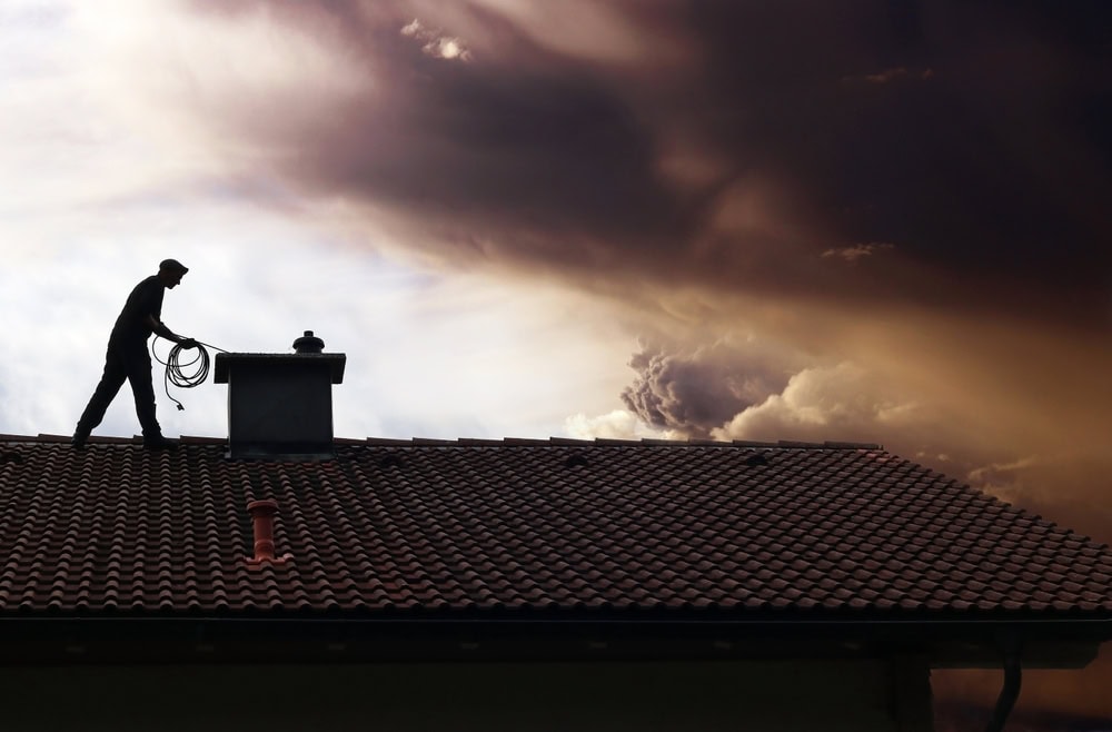 A silhouette of a person working on a tiled roof near a chimney, holding a coiled rope, captures the essence of chimney repair Norfolk and Plymouth County under dramatic, dark clouds in the sky behind them.