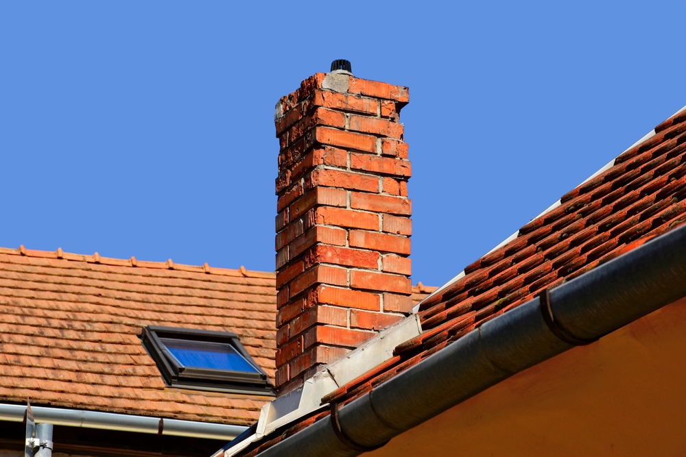 A red brick chimney rises above a tiled roof with a skylight window under a clear blue sky. A metal gutter runs along the edge, perfect for homes needing chimney repair in Norfolk and Plymouth County.