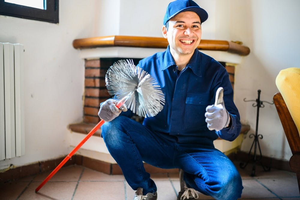 A smiling worker in blue overalls and cap crouches indoors, holding a chimney cleaning brush and giving a thumbs-up gesture—ready for expert chimney repair Norfolk and Plymouth County, MA.