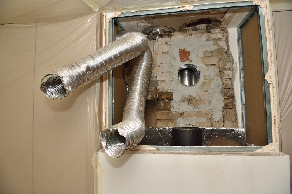 Two flexible metal ventilation ducts extend from an opening in a wall with exposed bricks and a circular vent—typical of chimney repair in Norfolk and Plymouth County, MA. The unfinished area is covered in plastic sheeting.