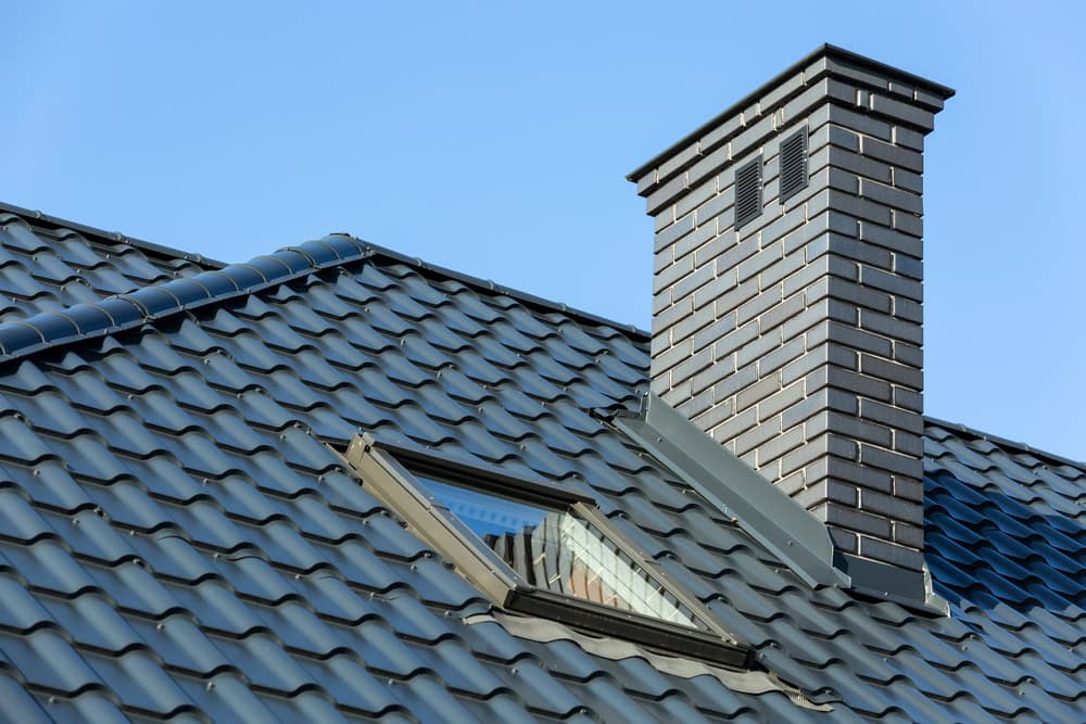 A close-up view of a house roof with dark blue metal tiles, a skylight window, and a brick chimney—highlighting expert chimney restoration Norfolk and Plymouth County—set against a clear blue MA sky.