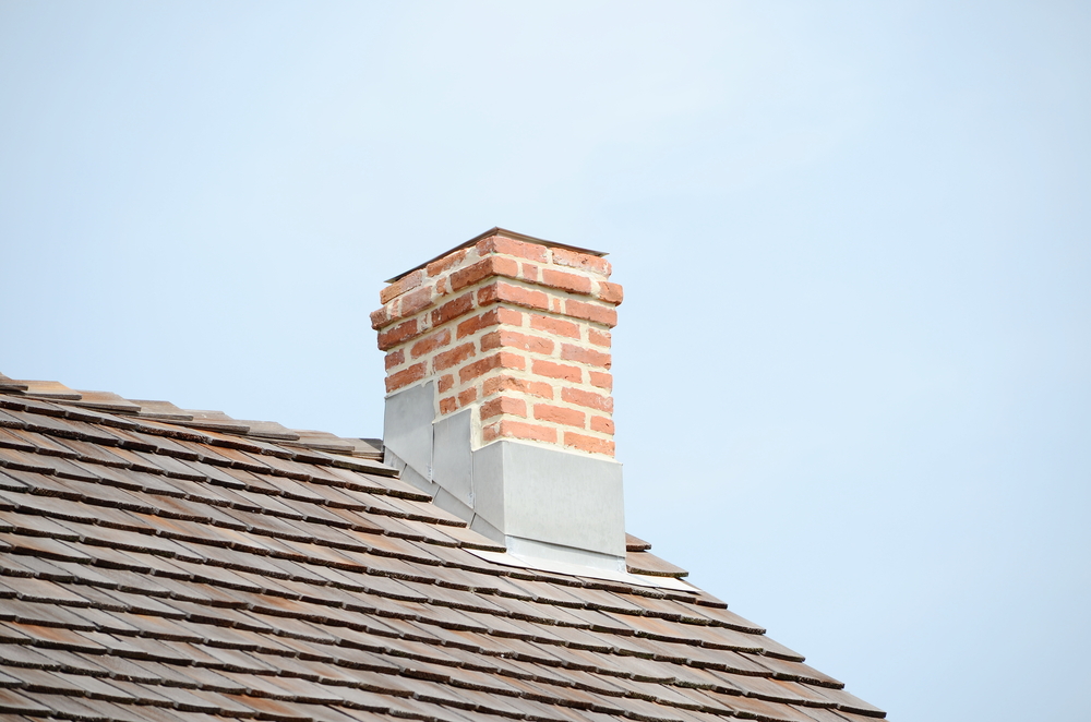 A red brick chimney with metal flashing extends from a sloped roof covered in brown shingles, set against a clear, pale blue sky—an ideal candidate for chimney restoration in Norfolk and Plymouth County, MA.