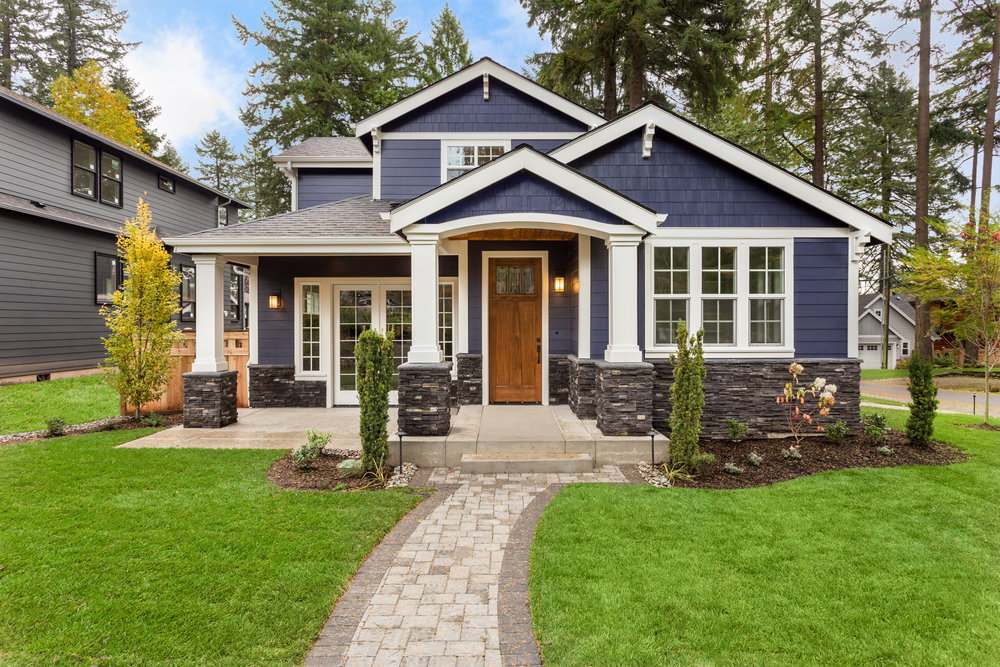 A modern two-story house in MA with navy blue siding, white trim, a wooden front door, and stone accents. It features a covered porch, manicured lawn, landscaping, a stone pathway to the entrance, and expert chimney repair Norfolk and Plymouth County, MA.