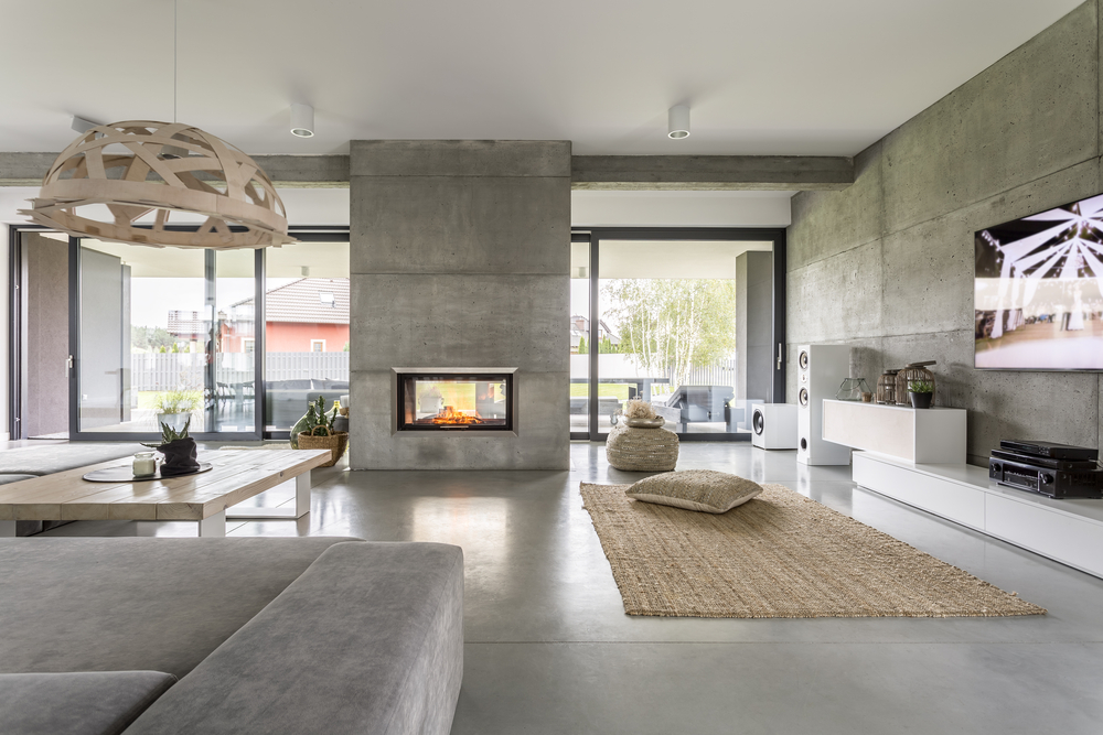 Modern living room with concrete walls and floors, a fireplace featuring chimney restoration Norfolk and Plymouth County, MA, large windows, gray sectional sofa, wooden table, woven rug, floor cushions, and a TV on a low white cabinet.