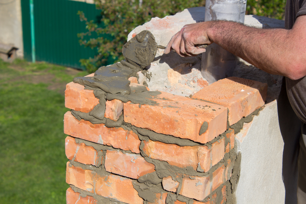 A person uses a trowel to apply wet mortar between orange bricks while working on chimney repair Norfolk and Plymouth County, MA, constructing or restoring a brick wall outdoors on a sunny day.