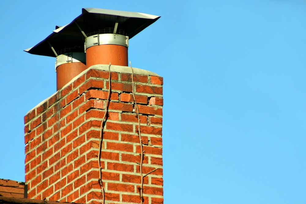 A brick chimney with two metal flues and a metal cap shows a large vertical crack running down the bricks, against a clear blue sky—an issue that may require chimney repair in Norfolk and Plymouth County, MA.