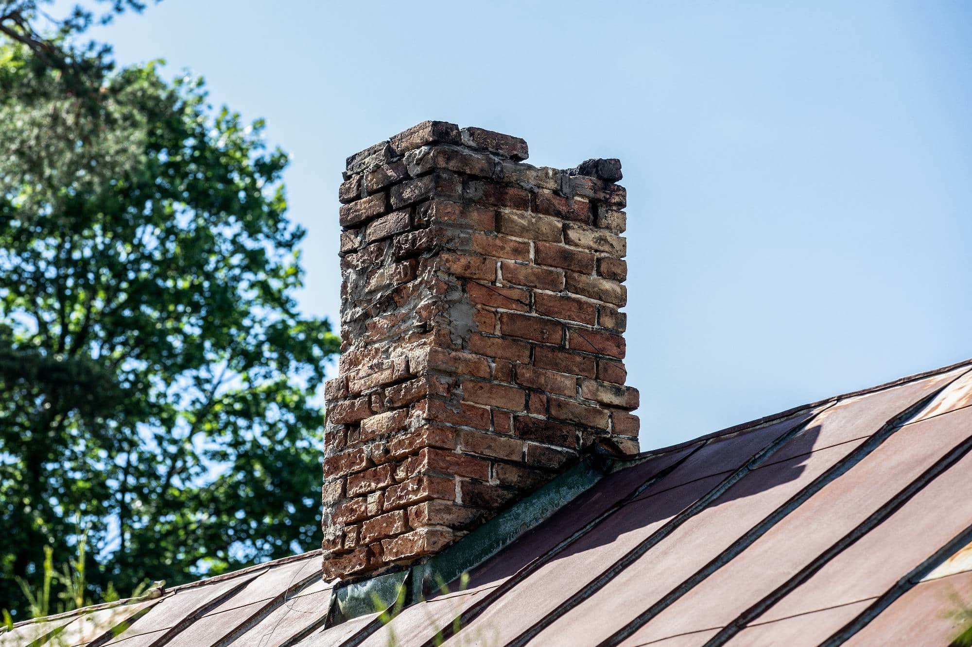 A weathered, crumbling brick chimney rises from a rusted metal roof, surrounded by green trees and set against a clear blue sky.