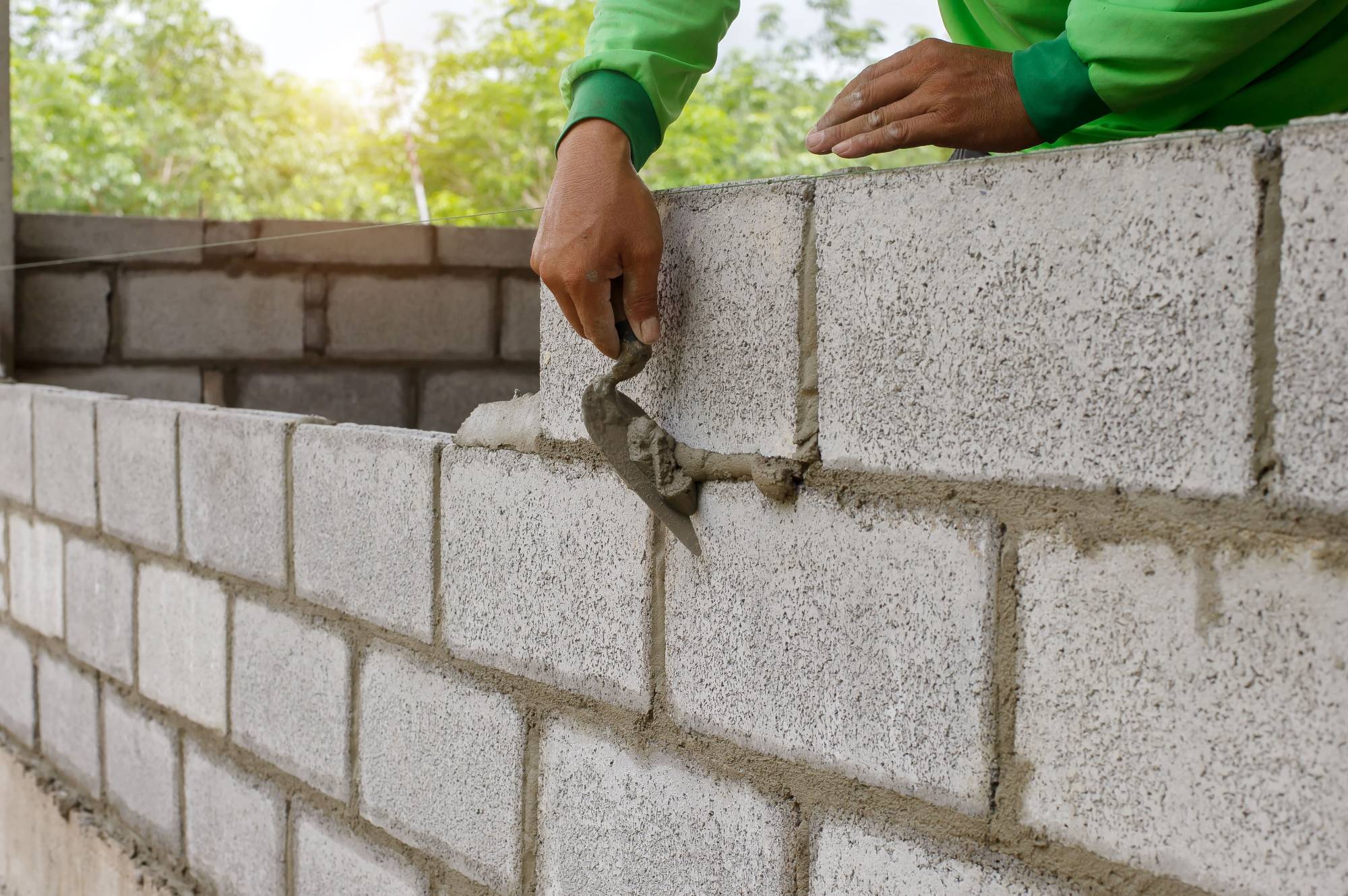 A person in a green shirt uses a trowel to spread mortar between concrete blocks while constructing a wall outdoors. Sunlight and greenery are visible in the background.