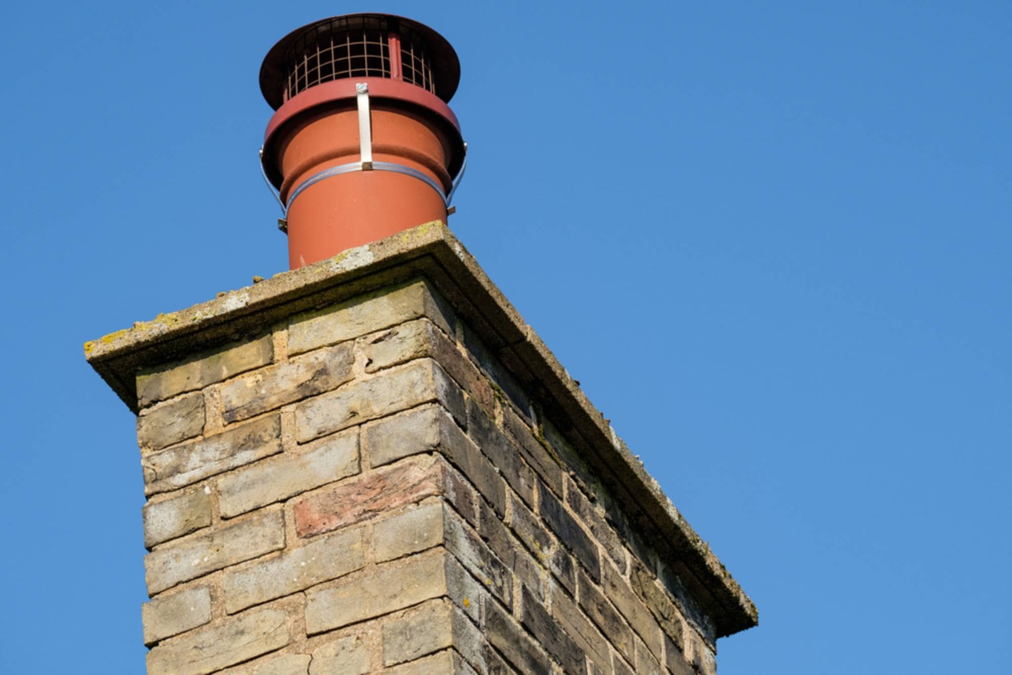 A close-up of a brick chimney with a red metal cap against a clear blue sky.