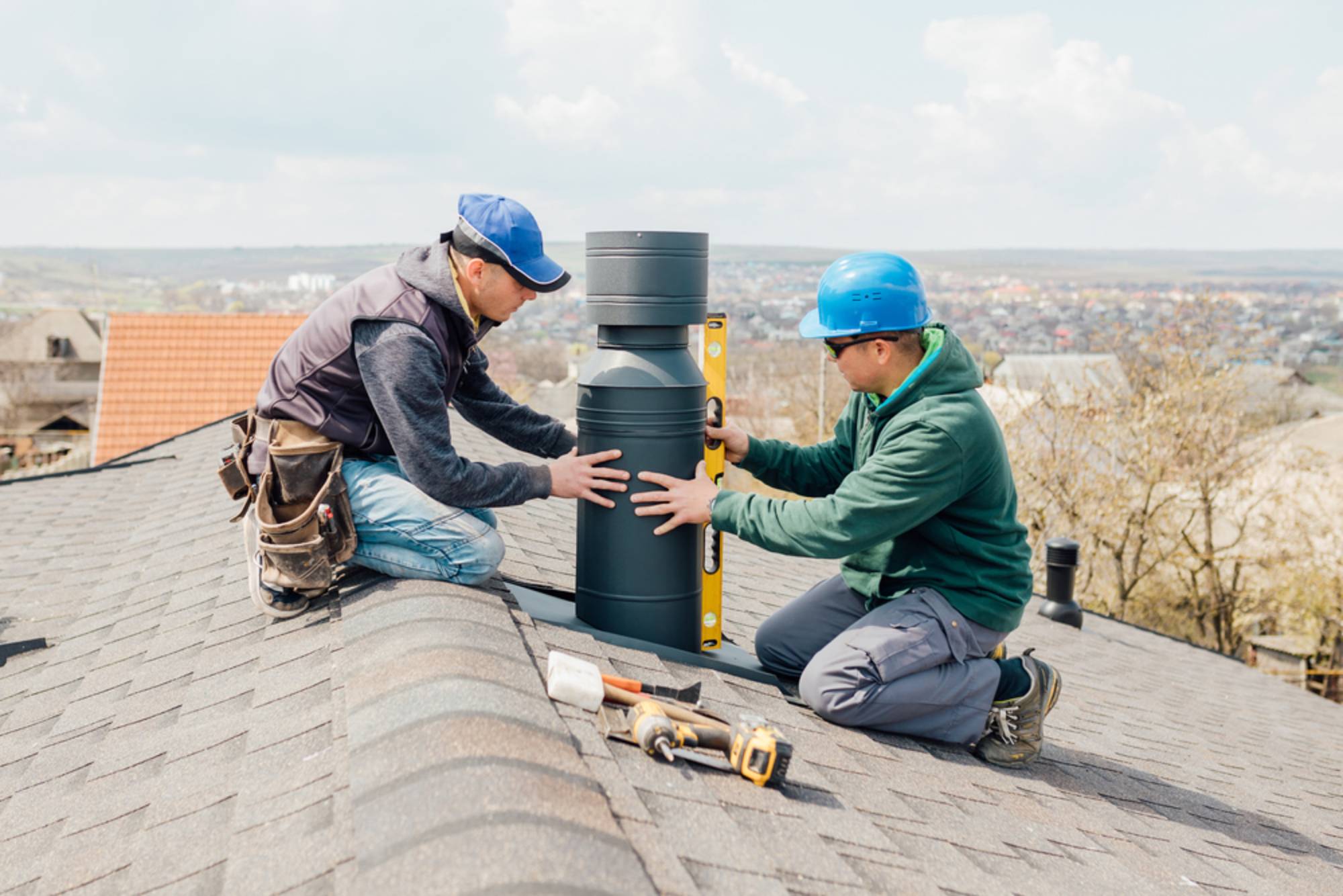 Two workers in safety gear install a metal chimney pipe on a shingled roof; one uses a level tool to ensure alignment, while several tools lie beside them. A townscape is visible in the background.