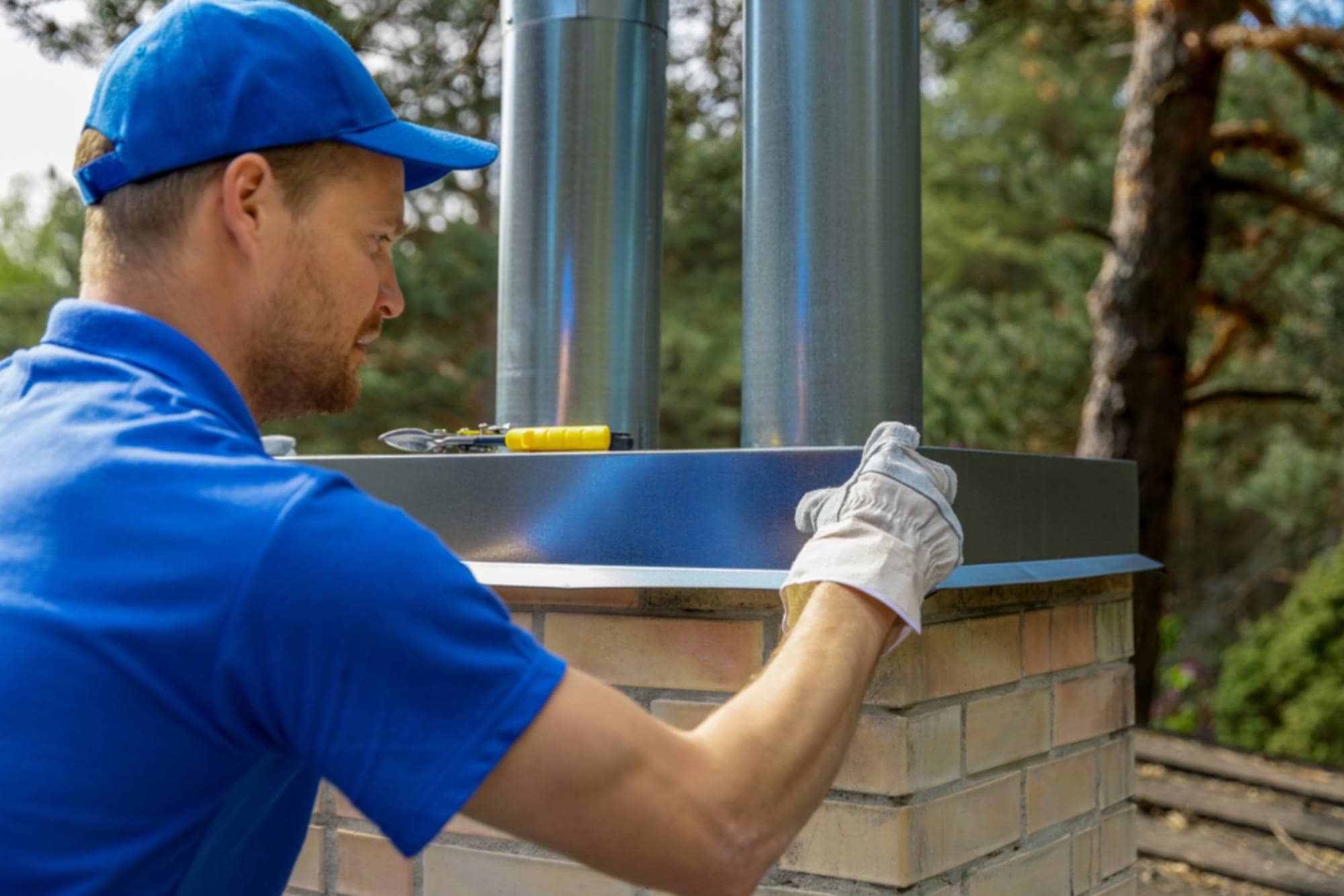 A man wearing a blue shirt, cap, and protective gloves installs or repairs a metal chimney cap on a brick chimney outdoors, with tools and green trees visible in the background.