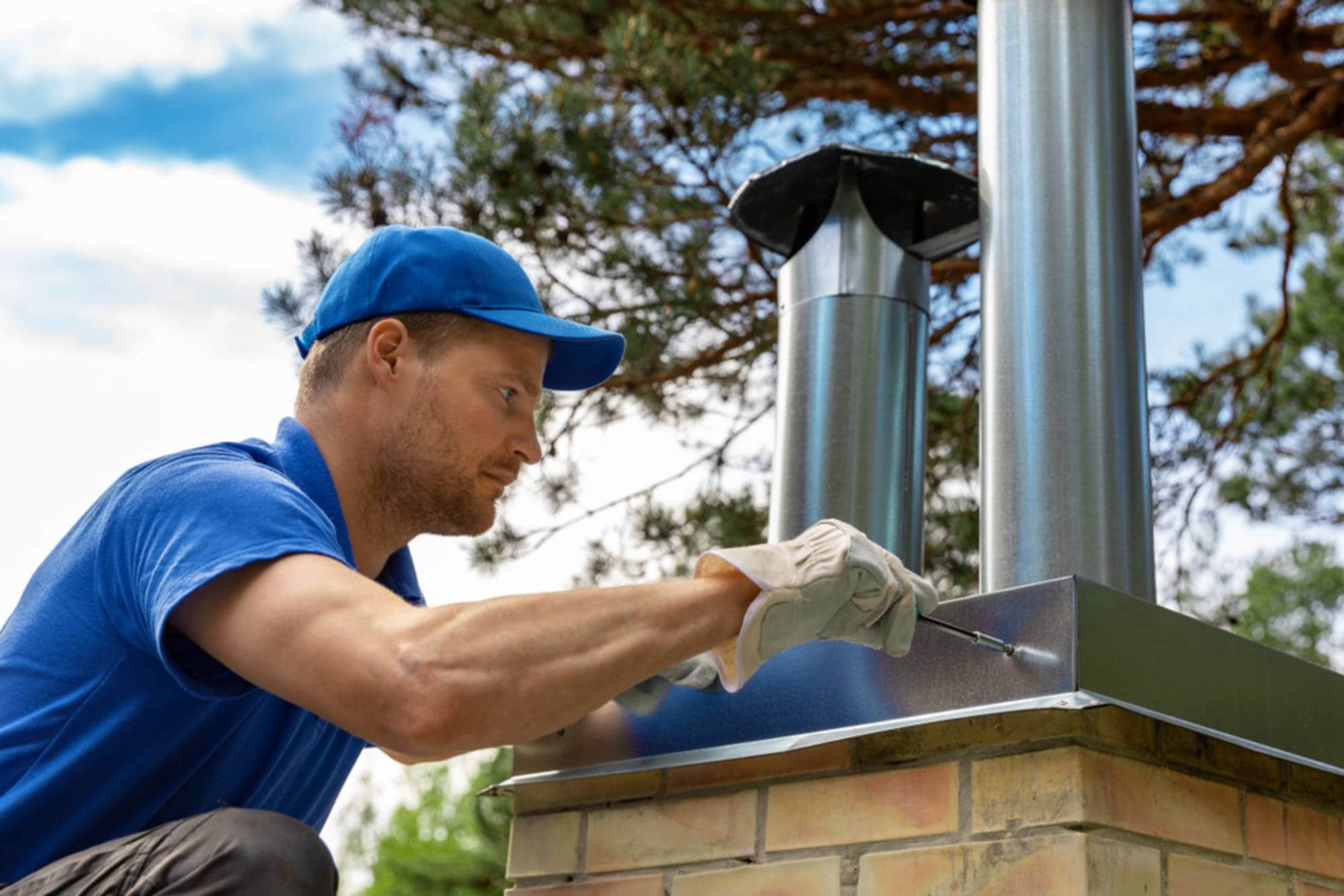 A person in a blue shirt and cap uses a screwdriver to install or repair a metal chimney on a brick structure outdoors, with trees and blue sky in the background.