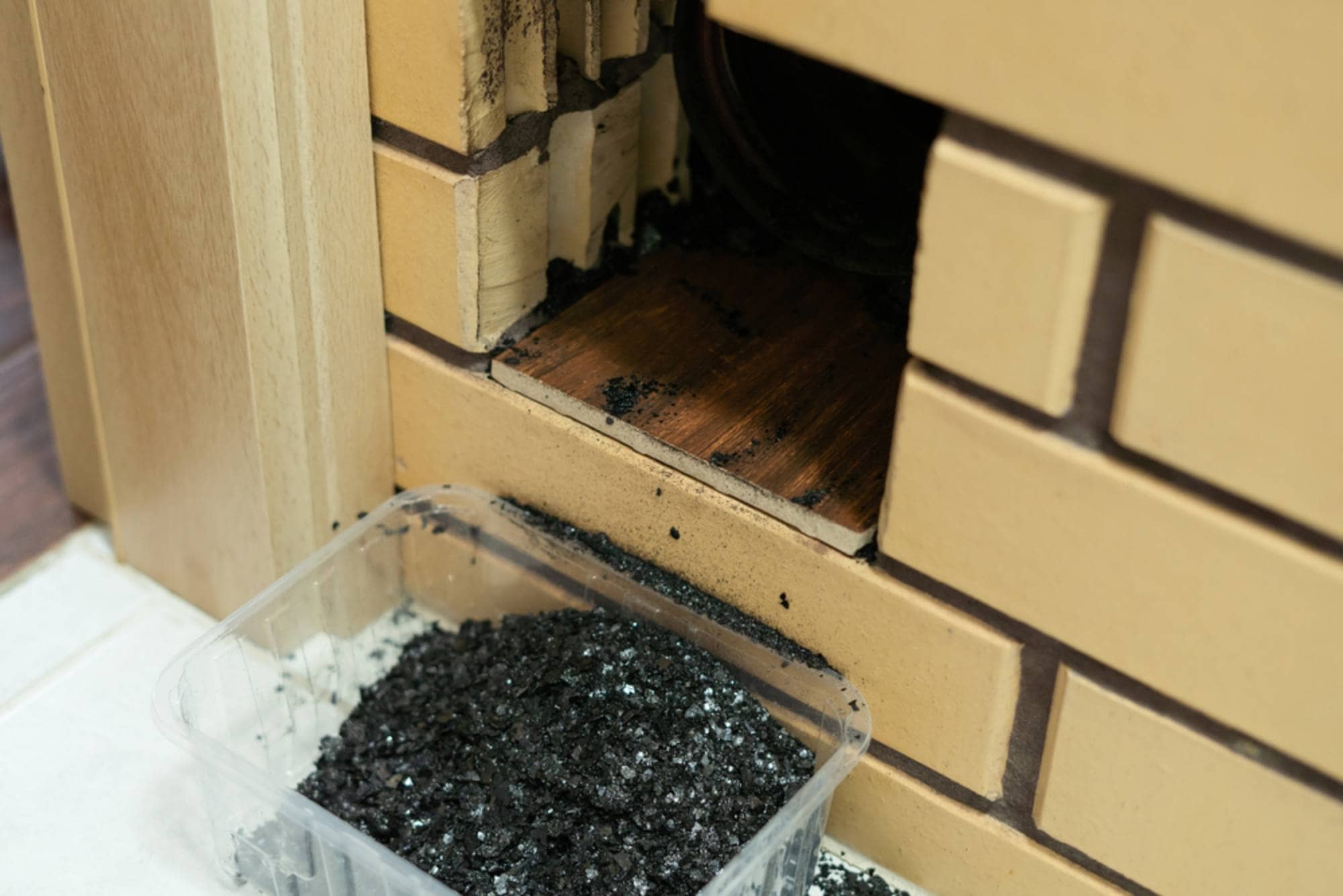 A close-up view of a brick fireplace interior shows an open vent exposing layers of dark soot and built-up ash along the inner walls. Beneath the opening, a plastic container has been positioned to catch falling debris, already filled with dark, powdery ash. The surrounding brickwork is stained from use, indicating that chimney or fireplace cleaning is actively underway. The image captures the gritty details of the cleaning process and highlights the accumulation typically found in a well-used fireplace.