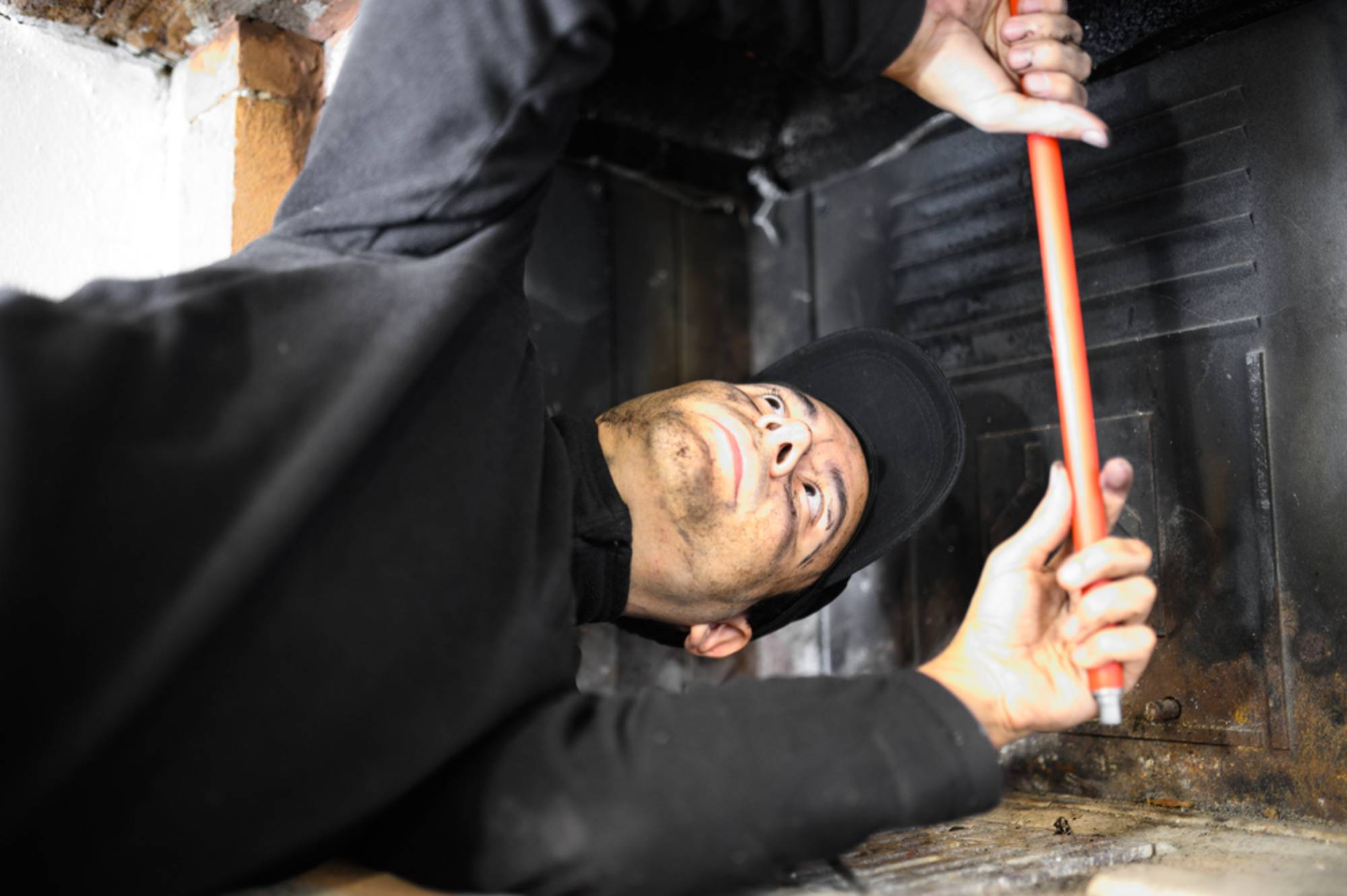 A person wearing a black cap and shirt uses an orange-handled tool to work inside a fireplace or chimney, looking upward while making repairs or maintenance.