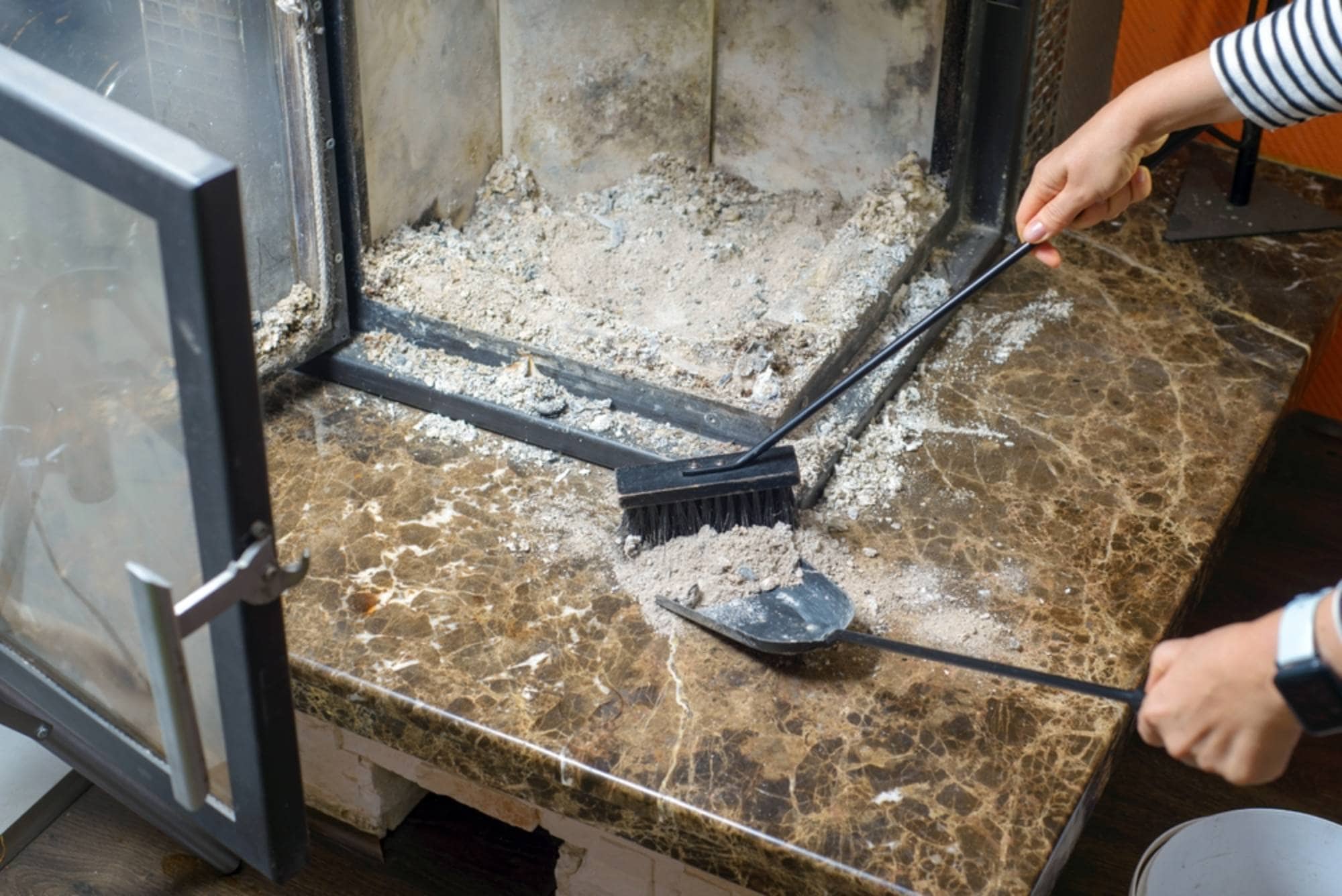 A person is cleaning a marble fireplace hearth using a handheld brush and dustpan, sweeping scattered gray ash into the pan. The fireplace doors are open, revealing a dark, soot-lined interior with more ash and residue visible inside. The marble surface is partially covered with fine ash particles, suggesting recent use of the fireplace. The person’s hands are visible mid-motion, emphasizing the act of careful cleaning and maintenance.