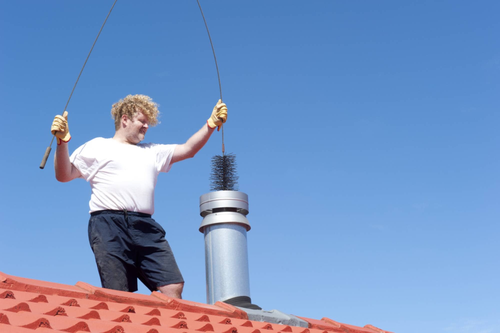 A person wearing gloves, a white t-shirt, and dark shorts stands on a red-tiled roof, holding a chimney brush above a metal chimney pipe under a clear blue sky.