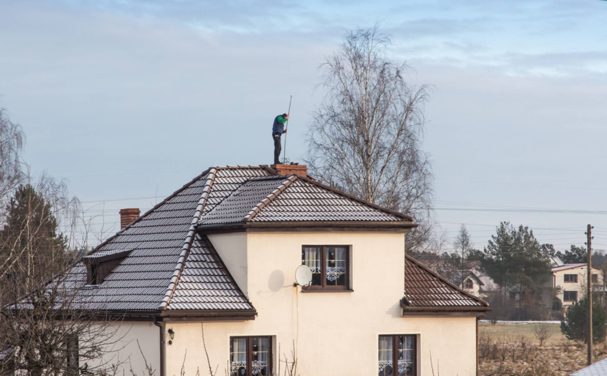 A person stands on the roof of a white house, cleaning or inspecting a chimney with a long tool. The house has brown roof tiles and is surrounded by trees and other houses in the background.