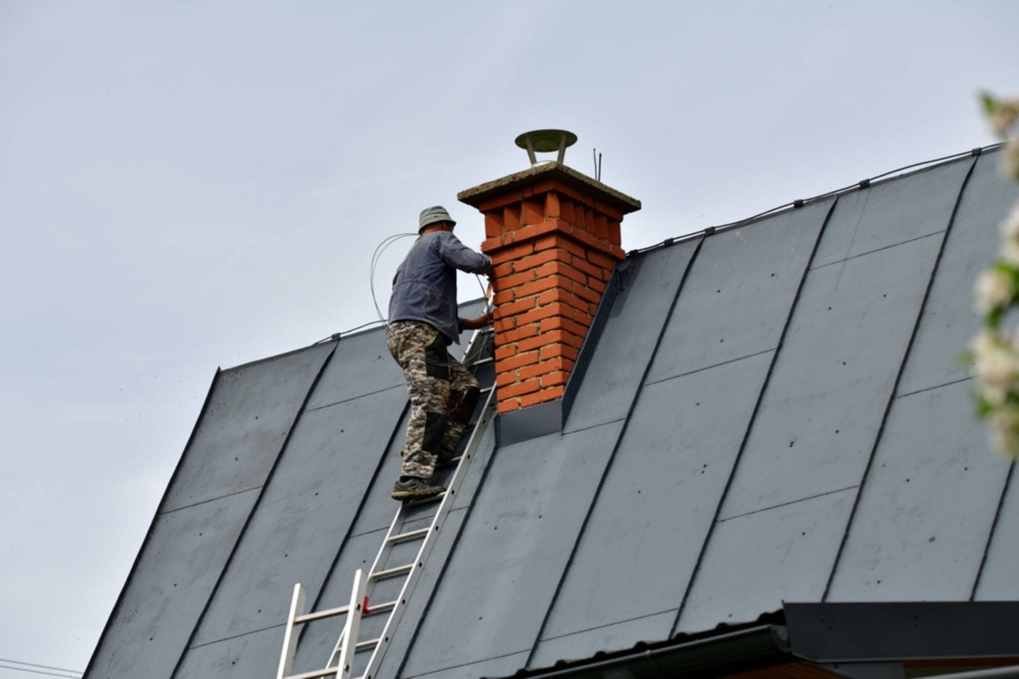 A person wearing camouflage pants and a hat stands on a ladder, working on a brick chimney atop a slanted, metal-roofed house under a clear sky.
